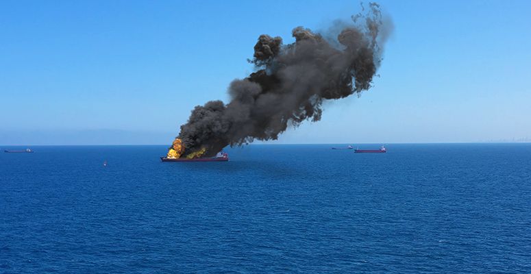 A wide-angle shot capturing a large vessel ablaze in the middle of the ocean, surrounded by other vessels.