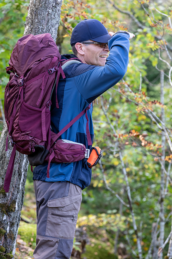 Hiker in active wear and hiking gear gazes across wooded area, equipped with Tron SA20 Locator Beacon attached to jacket