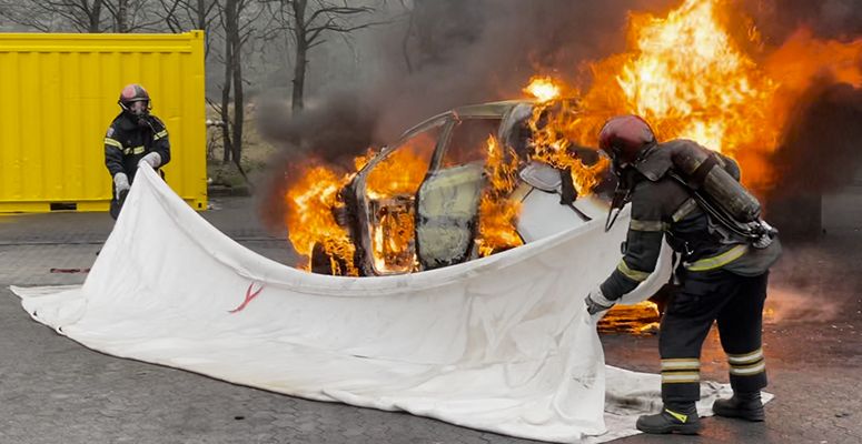 The Fire Isolator Blanket in action, deployed by two fully equipped firefighters as they extinguish a fire on a white car.